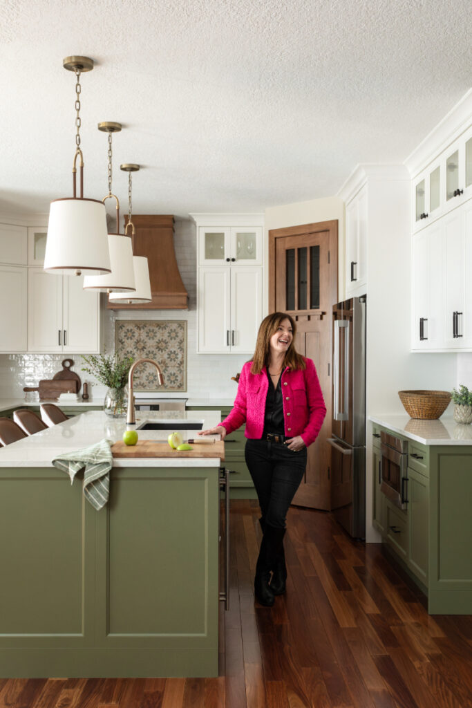 A project manager stands smiling in a remodeled kitchen with green cabinets, wood accents, and pendant lighting, showcasing the finished design.
