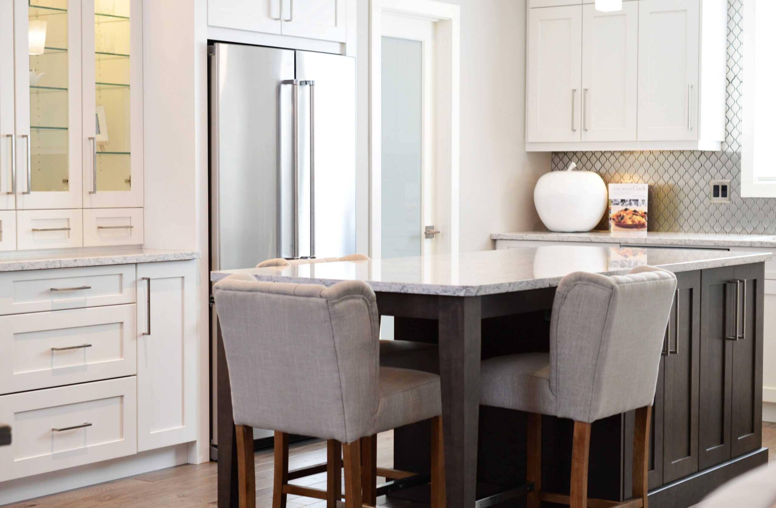 Modern kitchen with a marble island, two upholstered chairs, stainless steel fridge, white cabinets, and a decorative vase on the counter.