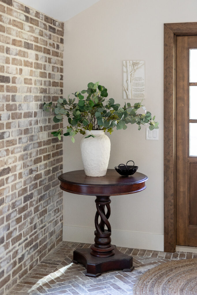 Entryway with brick wall, round wood pedestal table, and vase with greenery.