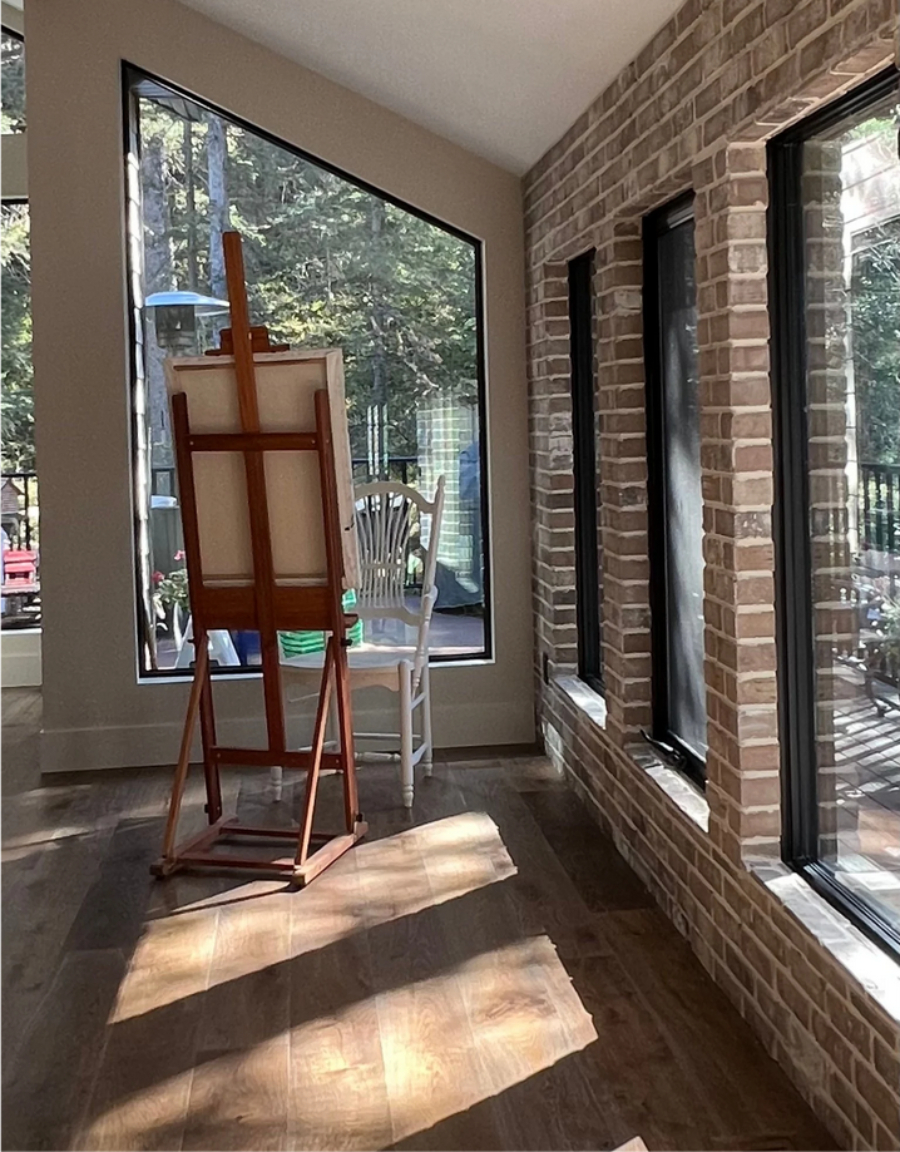 Sunlit studio with brick walls, tall windows, warm flooring, and an artist’s easel set in natural light.