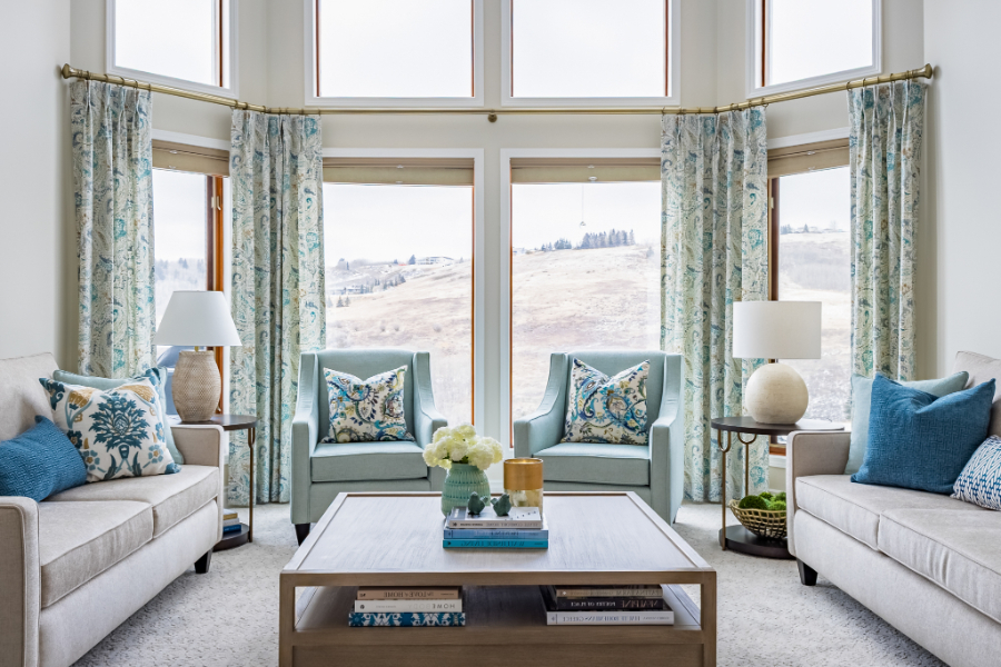Bright, inviting living room with large windows, soft neutrals, blue accents, and symmetrical seating arranged around a modern coffee table.