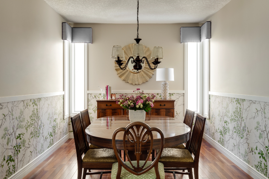 Traditional dining room with floral wainscoting, wood furniture, twin windows, and a central chandelier above a round table with a fresh flower arrangement.