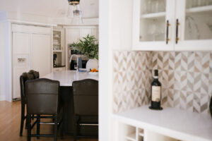 View of a bright, modern kitchen with patterned backsplash, white countertops, dark chairs, and open shelving.