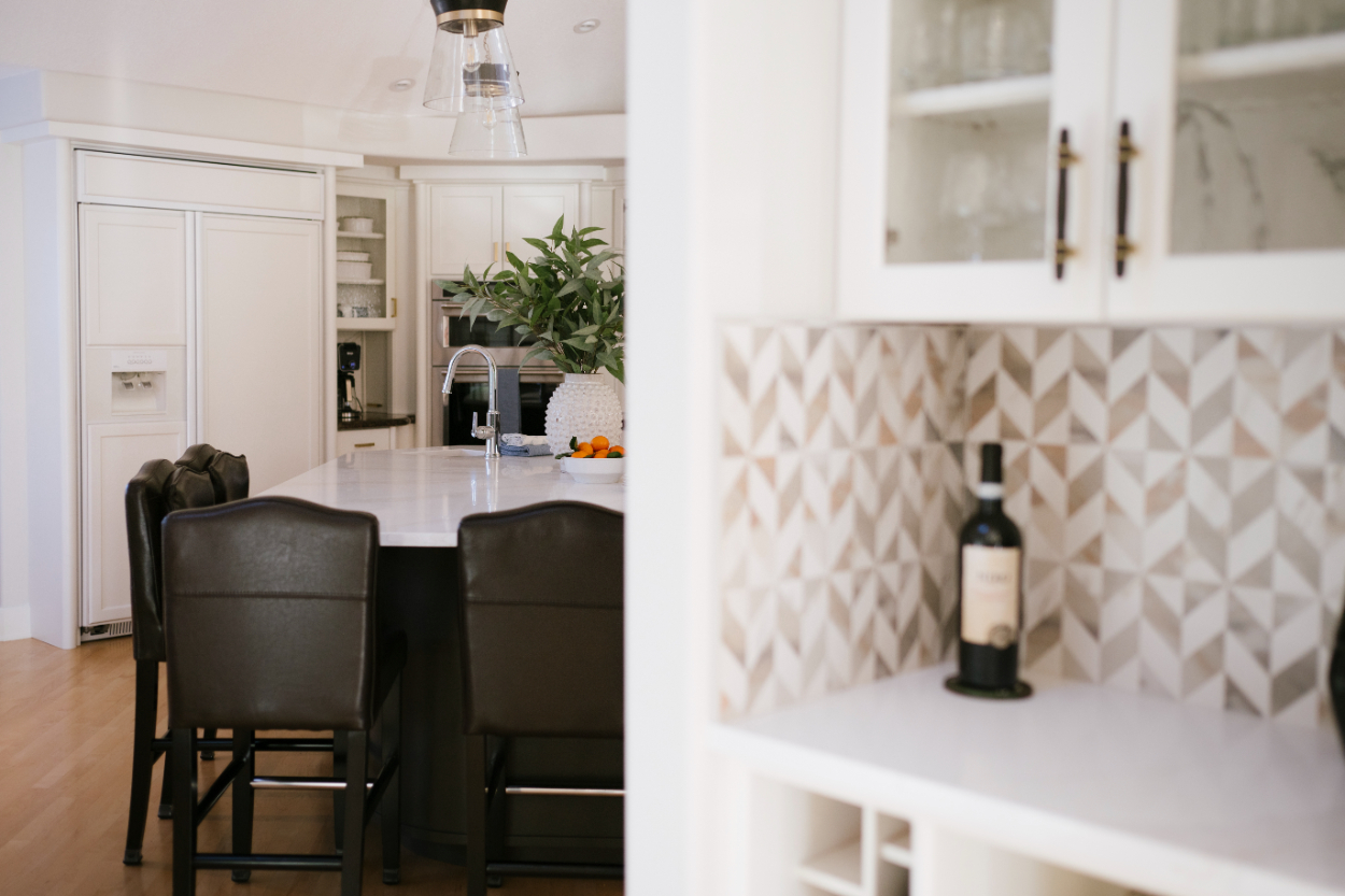 View of a bright, modern kitchen with patterned backsplash, white countertops, dark chairs, and open shelving.