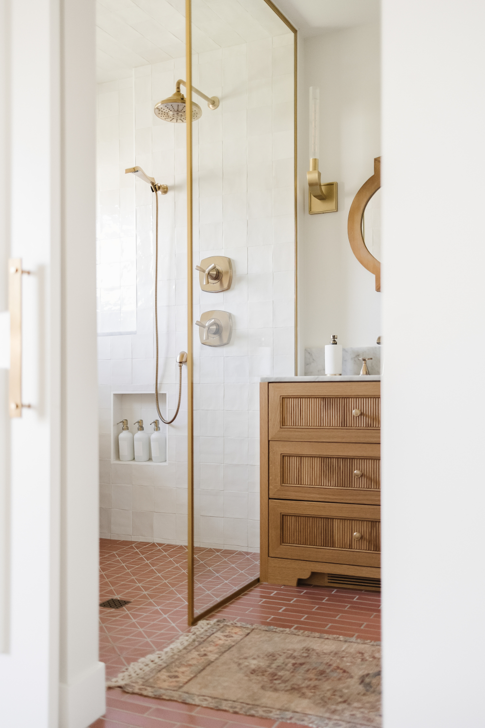 Modern bathroom with glass shower, brass fixtures, white textured tiles, and terracotta floor. Wooden vanity with marble top and round mirror sits beside a vintage rug and three white bottles in a shower niche.