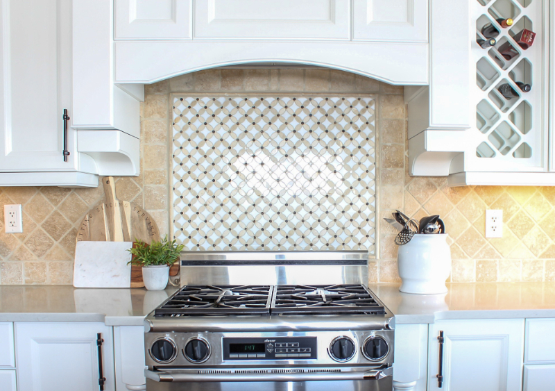 Bright kitchen stove area with white cabinets, beige tile backsplash, patterned inset above stainless range, and countertop decor including cutting boards, plant, and utensil jar.