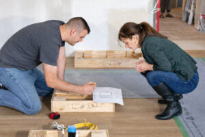 Two people kneel on a floor reviewing a construction plan, marking measurements on wood framing pieces during a home renovation project.