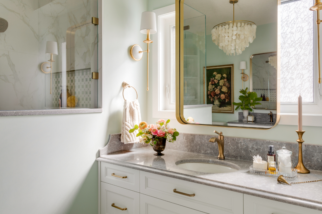White bathroom vanity with grey stone countertop, brass fixtures, gold-framed mirror, capiz shell chandelier, and sage green walls in a Room Service Interiors bathroom renovation.