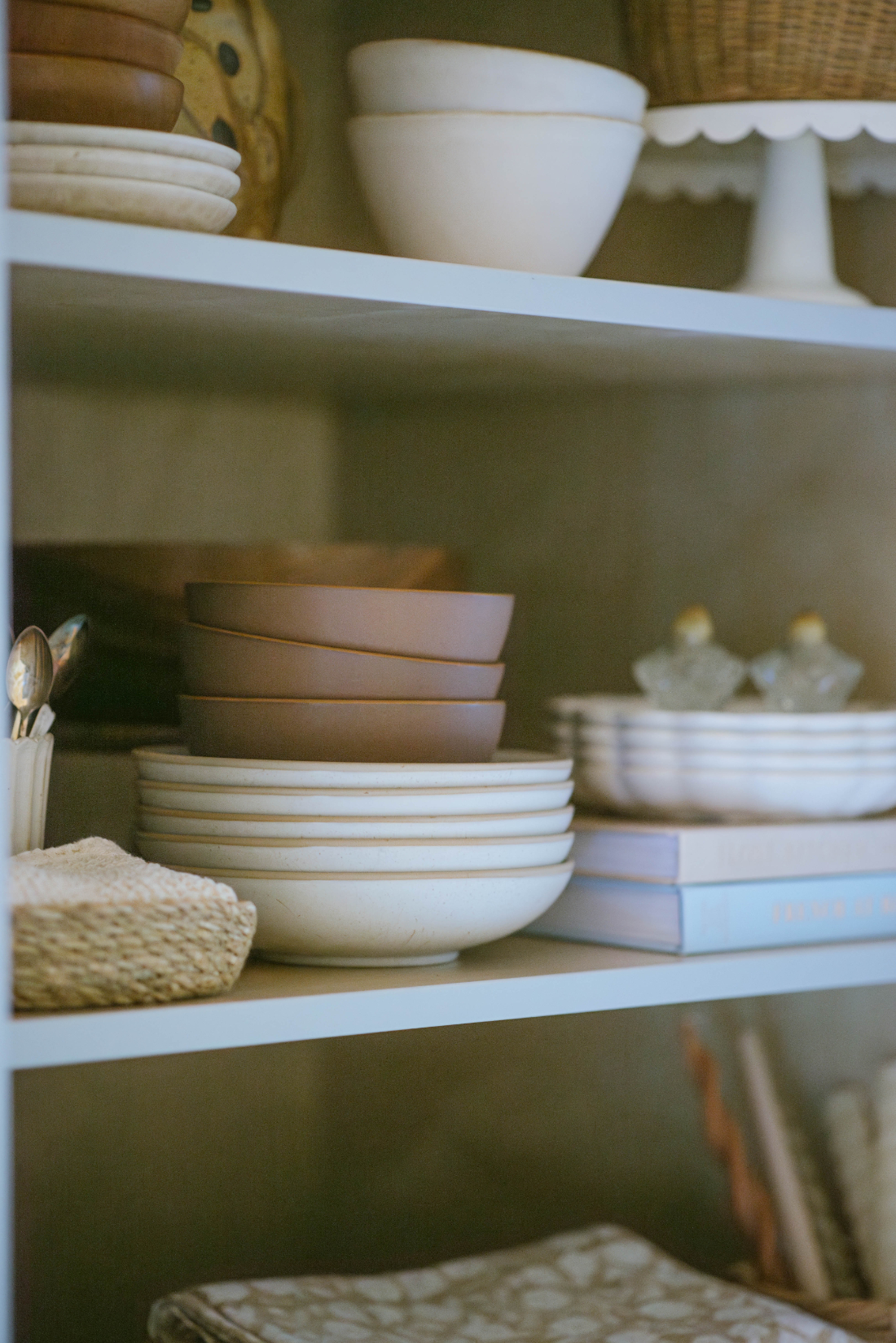Styled open shelving with neutral ceramic bowls, stacked plates, woven textures, and soft earthy tones in a cozy kitchen space.