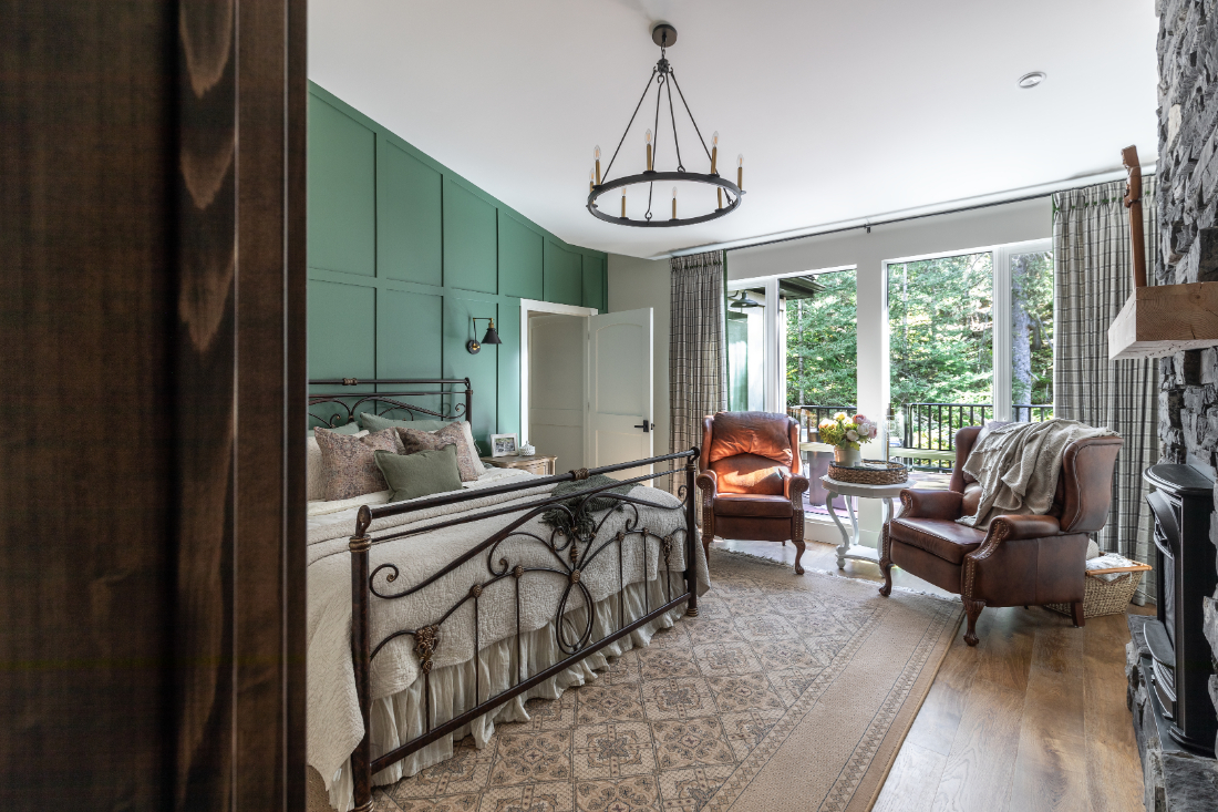 Cozy mountain-style bedroom with green accent wall, iron bed frame, leather chairs, and forest views through large windows.