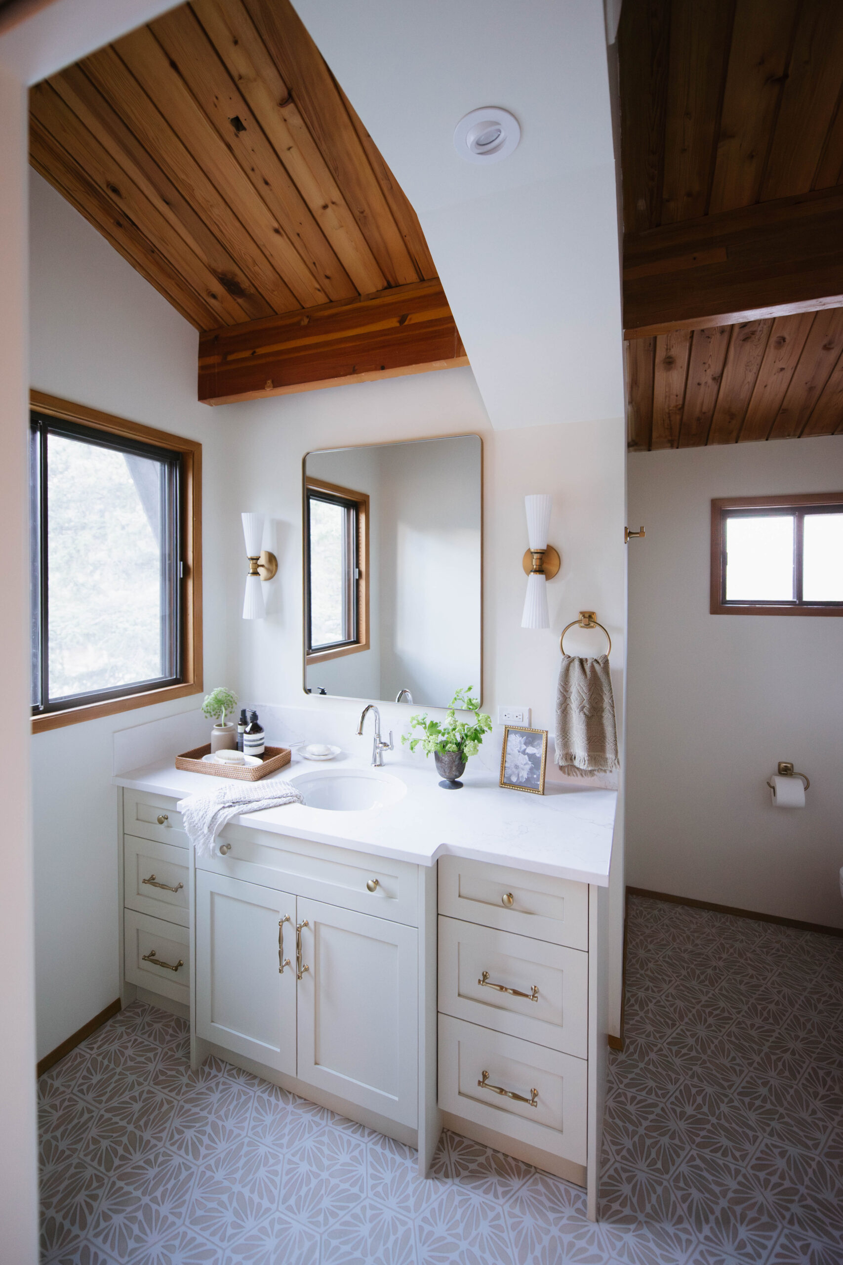 Bathroom renovation by Room Service Interiors with a custom vanity, large mirror, and vaulted wood ceiling. Light finishes, reflective surfaces, and efficient storage demonstrate ideas for small spaces that feel bright and spacious.
